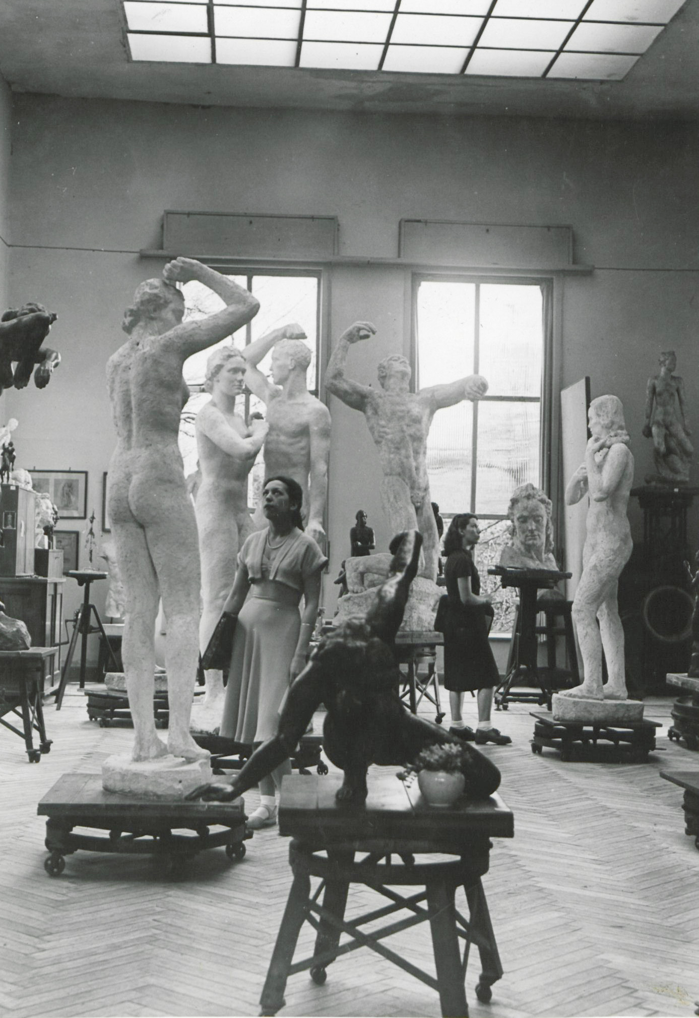 Schwarz-weiß-Foto von Skulpturen in einem hohen Raum. In der Mitte des Raums steht eine Frau, die eine der dargestellten Figuren betrachtet.