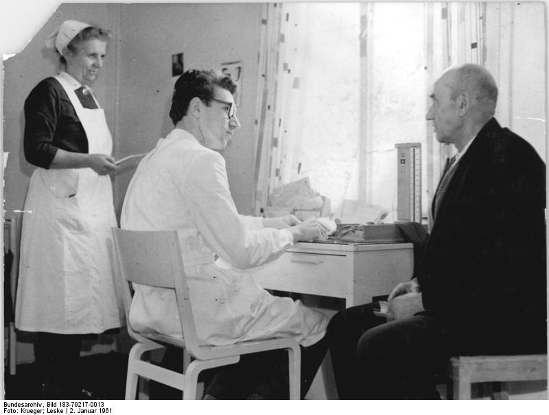 Das Foto zeigt zwei Männer, davon einer Arzt und einer Patient, beide sitzend und stehend daneben eine Krankenschwester in einem Zimmer.