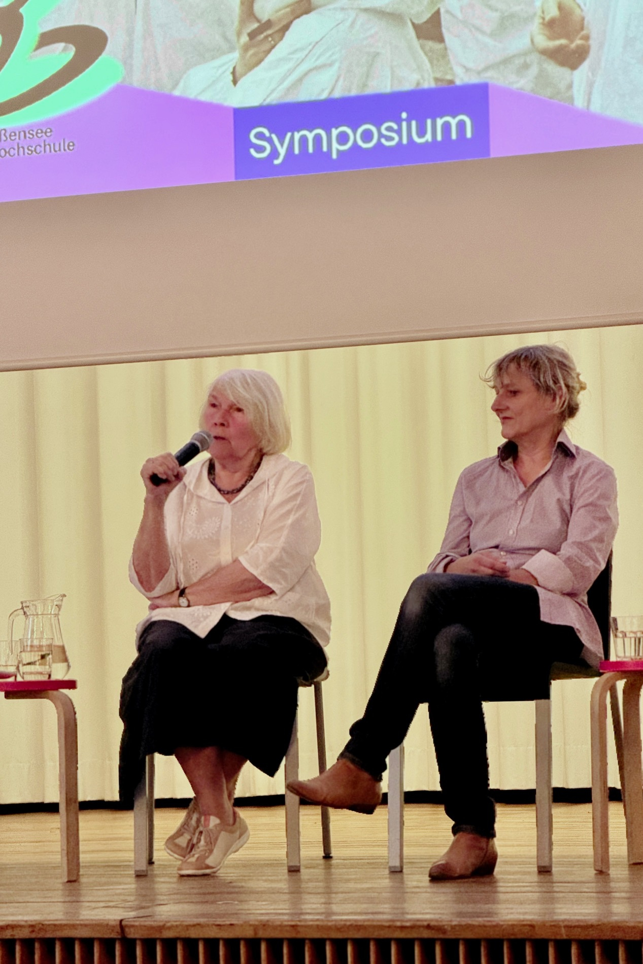 Jutta Wachowiak (Schauspielerin) links mit Mikrofon in der Hand auf dem Podium in der Aula der KHB sitzend. Neben ihr rechts Inken Reinert, Künstlerin und Absolventin der KHB.