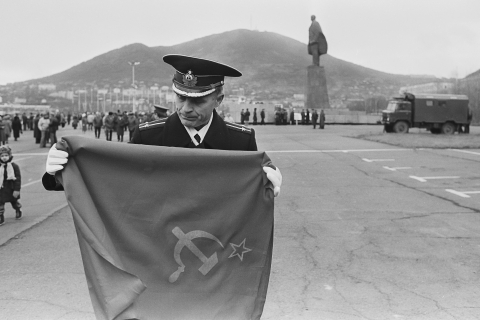 Auf einem großen Platz faltet ein Militärangehöriger eine Fahne der Sowjetunion zusammen, die er in den Händen hält. Im Hintergrund ist eine große Statue zu sehen, dahinter ein Hügel.