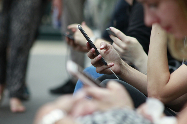 Teleaufnahme von Händen, die Smartphones bedienen. Der größte Teil des Bildes ist unscharf, nur in der Mitte ist eine scharfgestellte Hand mit roten Fingernägeln zu sehen, die ein Smartphone bedient.