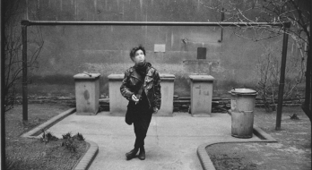 Schwarz-Weiß-Foto einer jungen Frau, die Zigarette rauchend im Innenhof eines Gebäudes steht. Im Hintergrund sind fünf Mülltonnen zu sehen, vier sind eckig, eine rund. Die Frau hat die Beine über Kreuz, exhaliert Rauch und schaut dabei schräg nach oben.