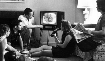 Bild einer amerikanischen Familie beim Fernsehsehen, ca 1958. Fünf Menschen sind um einen Fernseher gruppiert, teilweise liegen oder sitzen sie am Boden, eine Frau sitzt auf einem Sofa und hält eine Zeitung in der Hand. Foto: Evert F. Baumgardner