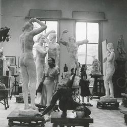 Schwarz-weiß-Foto von Skulpturen in einem hohen Raum. In der Mitte des Raums steht eine Frau, die eine der dargestellten Figuren betrachtet.