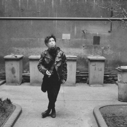 Schwarz-Weiß-Foto einer jungen Frau, die Zigarette rauchend im Innenhof eines Gebäudes steht. Im Hintergrund sind fünf Mülltonnen zu sehen, vier sind eckig, eine rund. Die Frau hat die Beine über Kreuz, exhaliert Rauch und schaut dabei schräg nach oben.