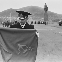 Auf einem großen Platz faltet ein Militärangehöriger eine Fahne der Sowjetunion zusammen, die er in den Händen hält. Im Hintergrund ist eine große Statue zu sehen, dahinter ein Hügel.